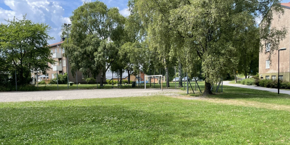 En grusbollplan med fotbollsplan intill en gräsmatta. Bolplanen är delvis inhägnad och bakom planen ses höga träd och flerbostadshus. Foto